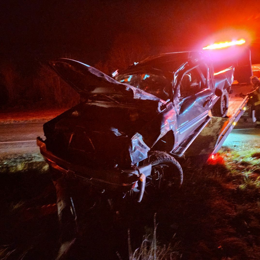 Severely damaged black car at night beside flashing red emergency lights on a wet roadside