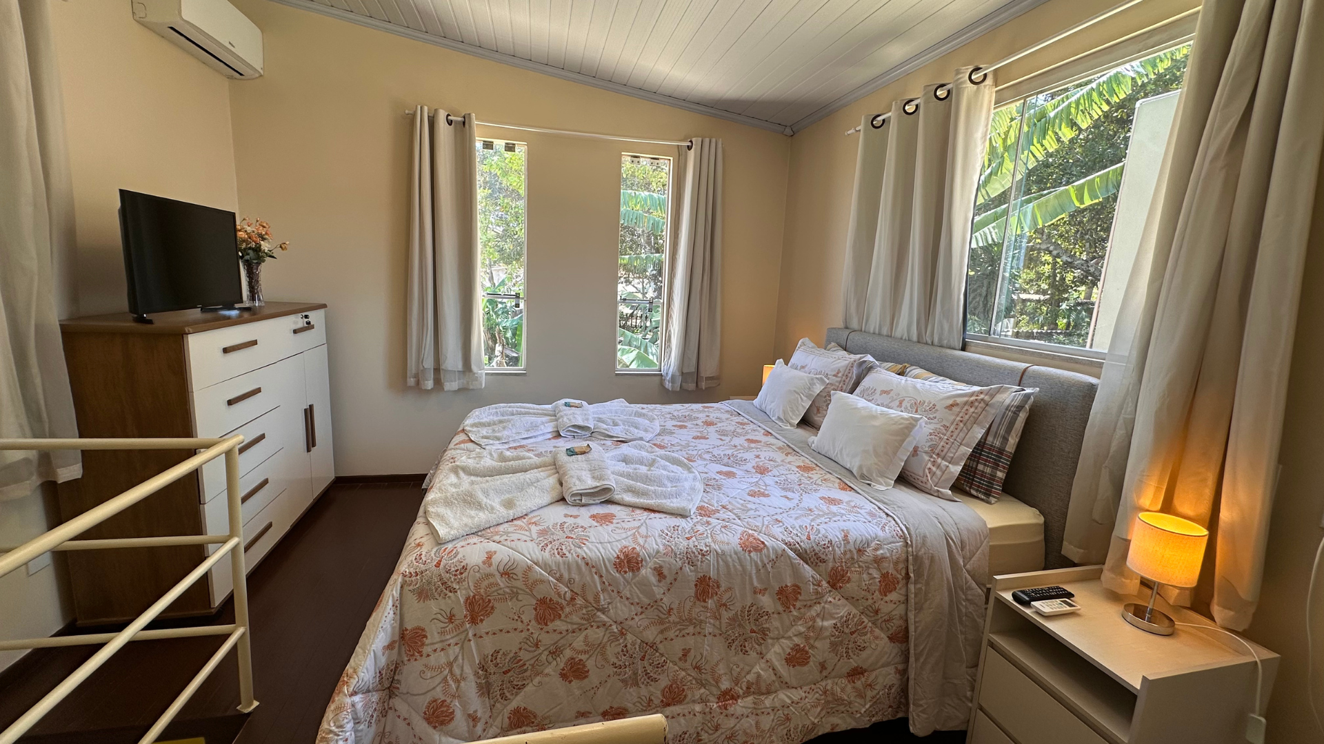 Quarto com cama de casal, cômoda, TV, duas janelas e mesa de cabeceira.
