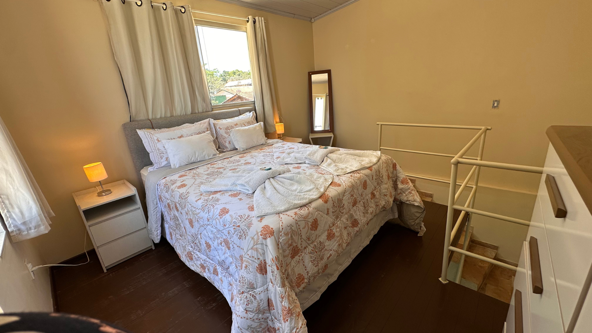 Quarto com cama, mesas de cabeceira, espelho de corpo inteiro e escada que leva ao andar inferior.