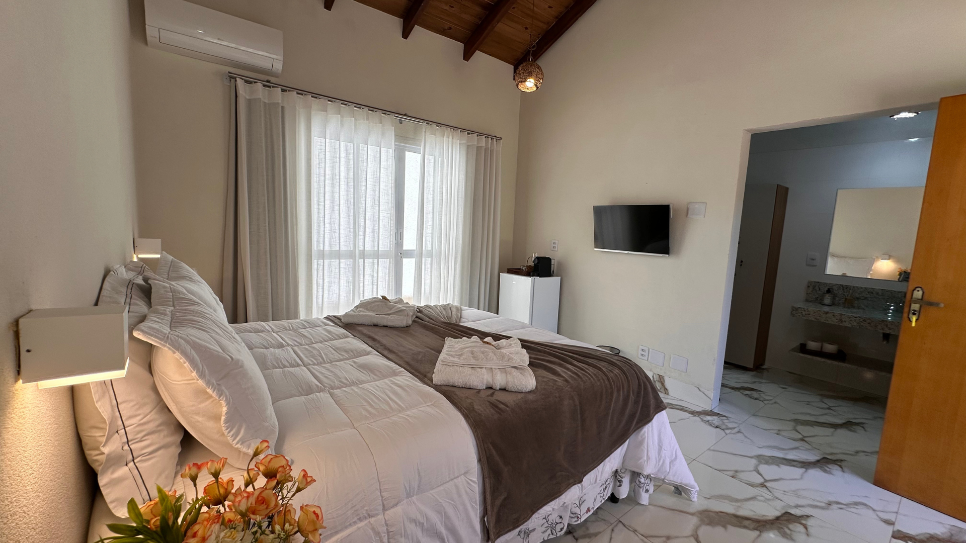 Quarto com cama grande, TV e banheiro privativo. Paredes em tons neutros, iluminação decorativa e janela.