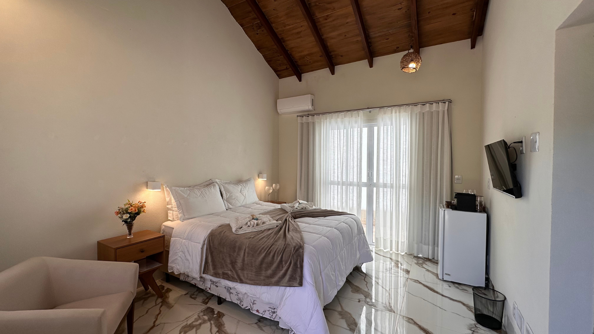 Quarto com cama, mesa de cabeceira, poltrona, cortinas e TV. Paredes bege e teto marrom.