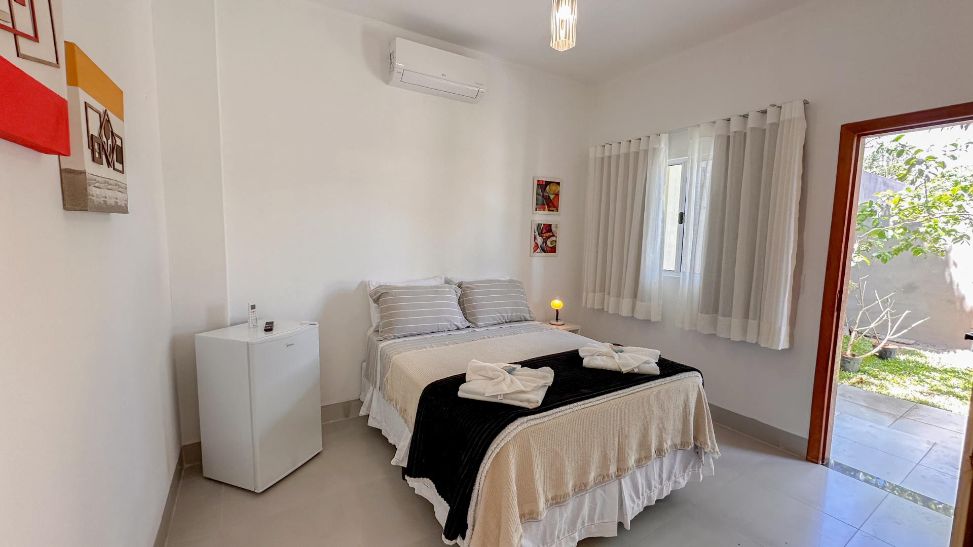 Quarto branco com cama, frigobar e porta aberta para um pátio.