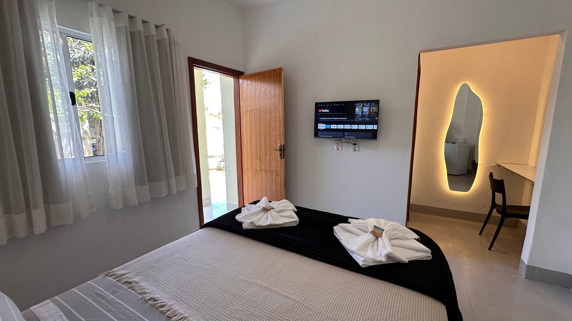Quarto com cama, TV, porta aberta e espelho decorativo com escrivaninha.