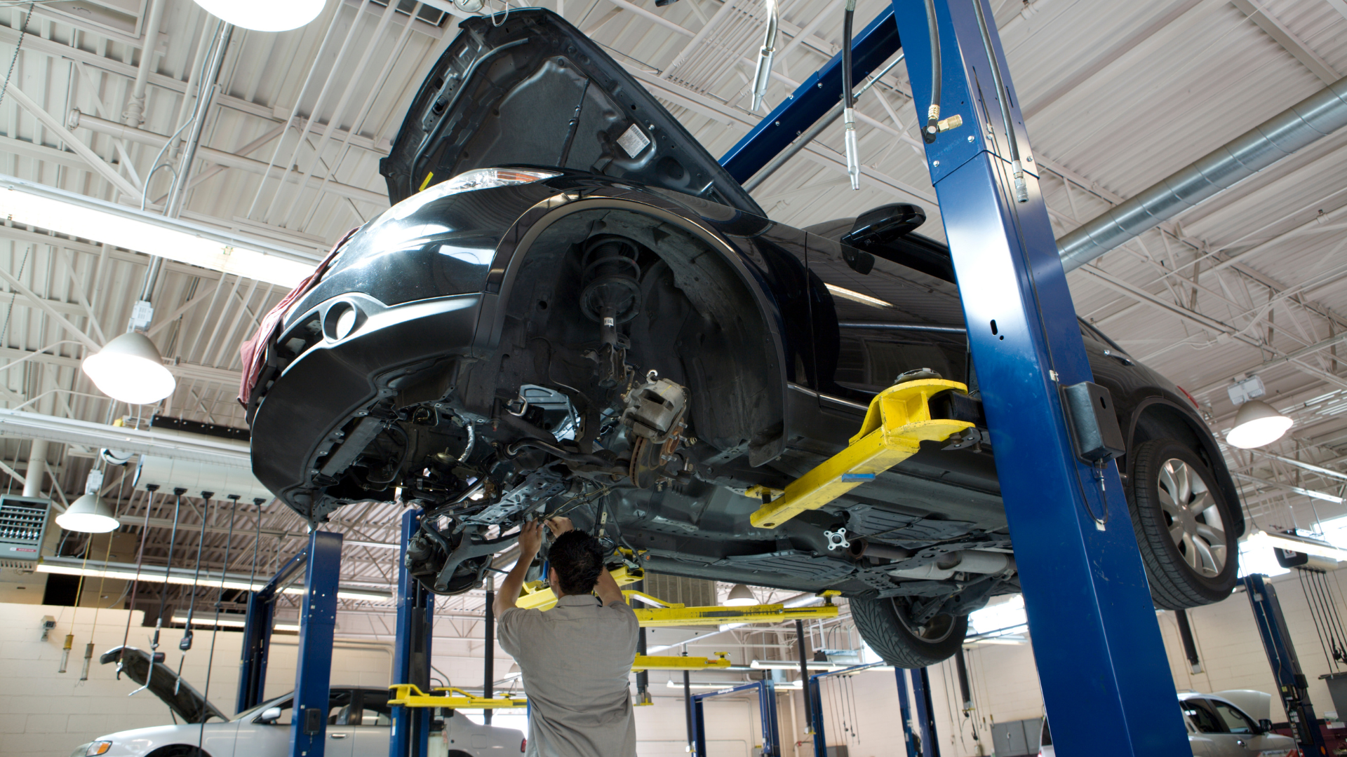 Black car lifted on a blue hydraulic hoist in an auto repair shop with a mechanic underneath