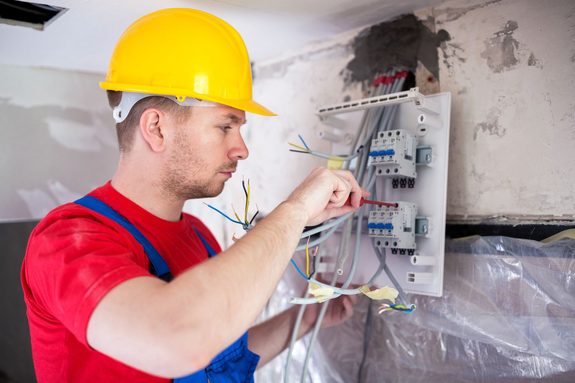 Electrician wearing a yellow hard hat performs electrical repair on an open circuit breaker panel.