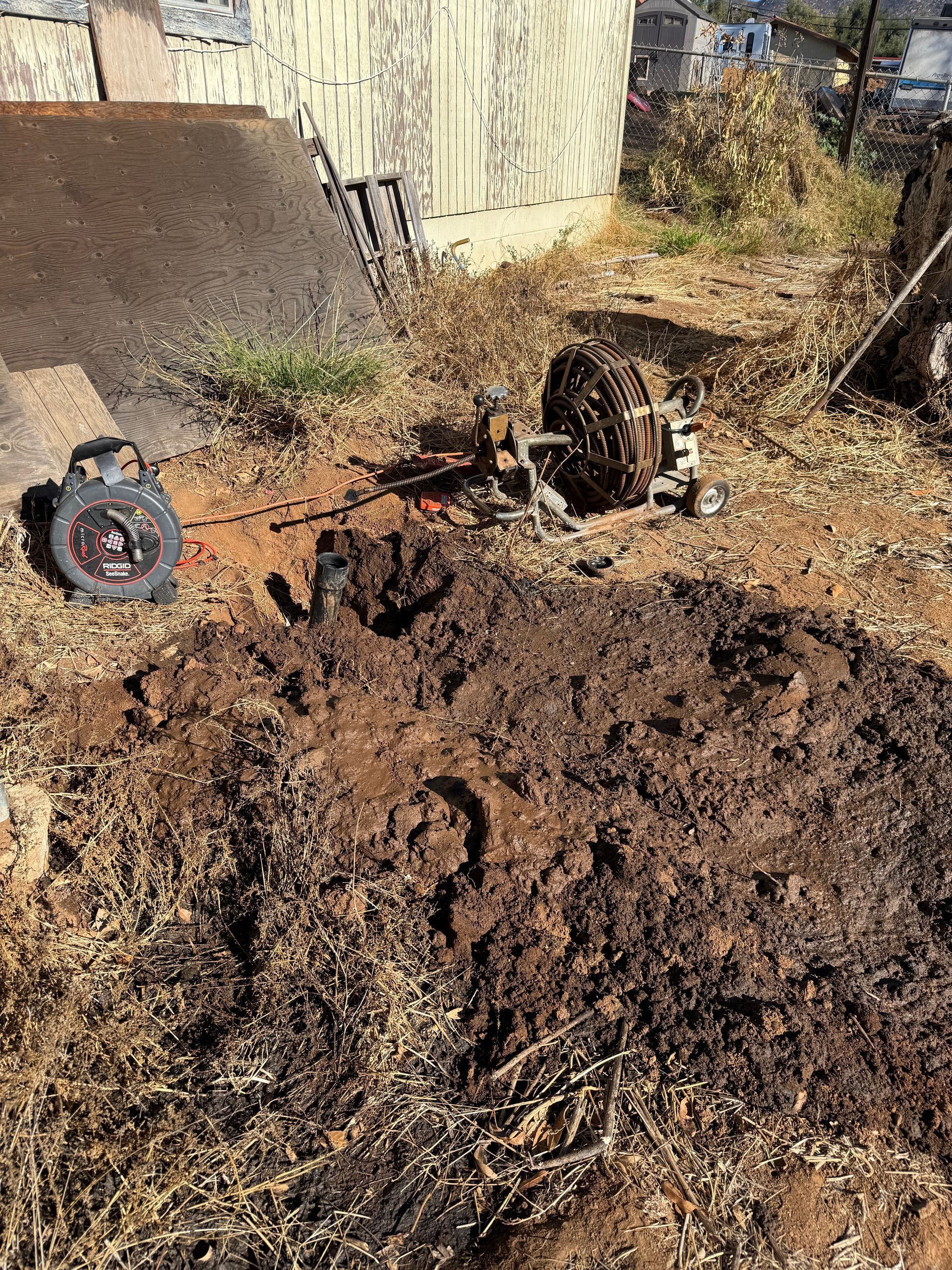 A metal spool and machinery equipment sit on a patch of loose, dark dirt near a concrete wall in an outdoor area.