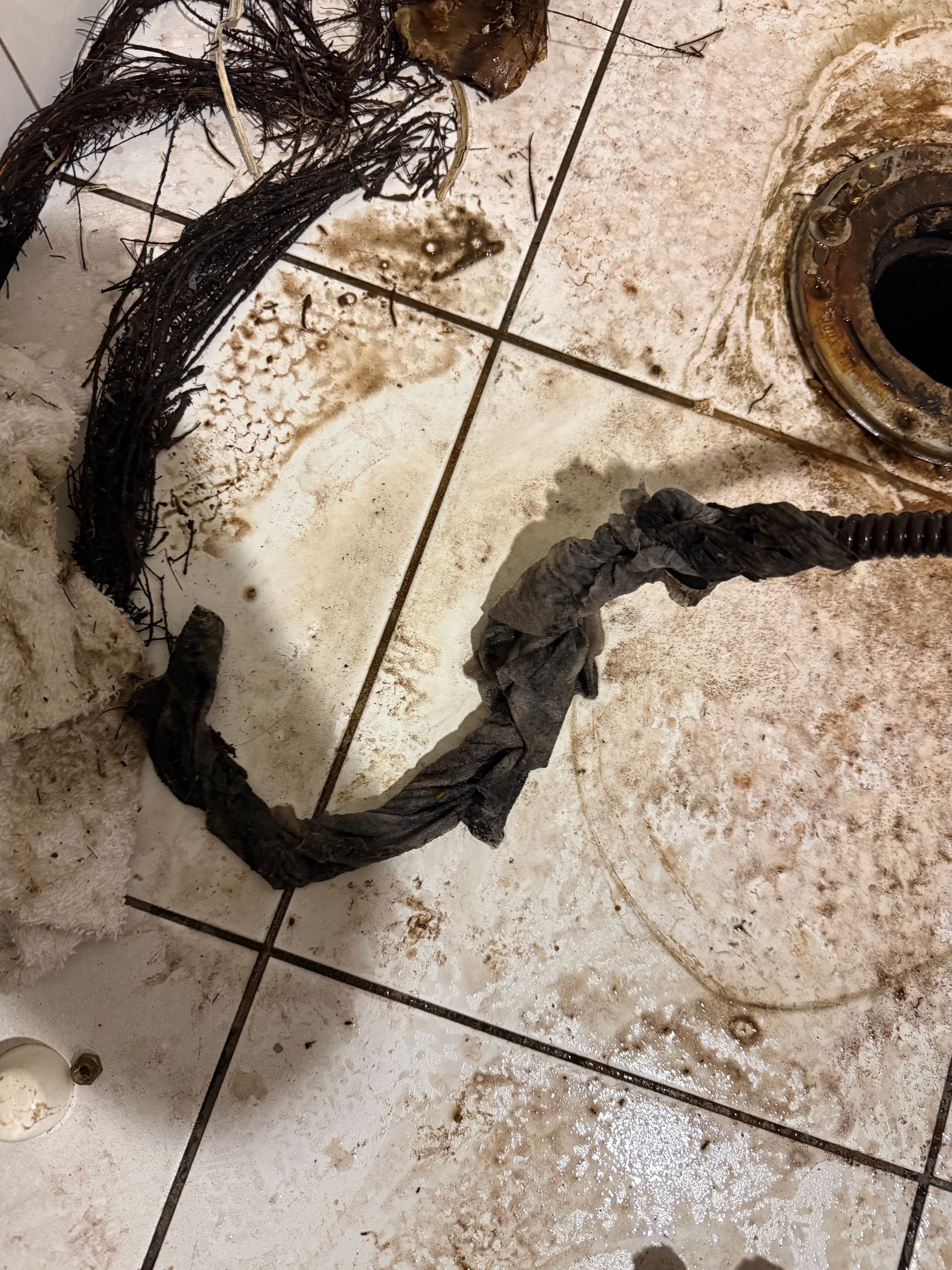 Debris from a drain, including hair and fabric, lying on a tiled bathroom floor next to a toilet drain pipe.