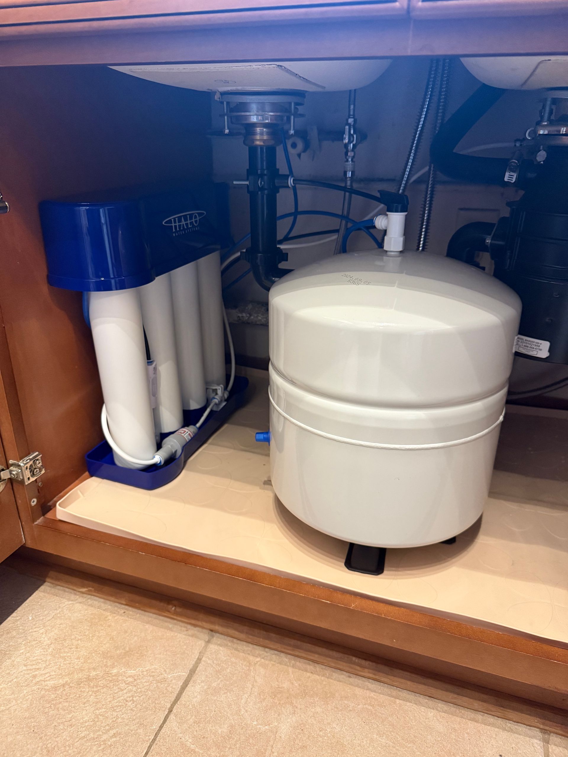 A reverse osmosis water filtration system with a white storage tank and blue filter manifold under a kitchen sink.