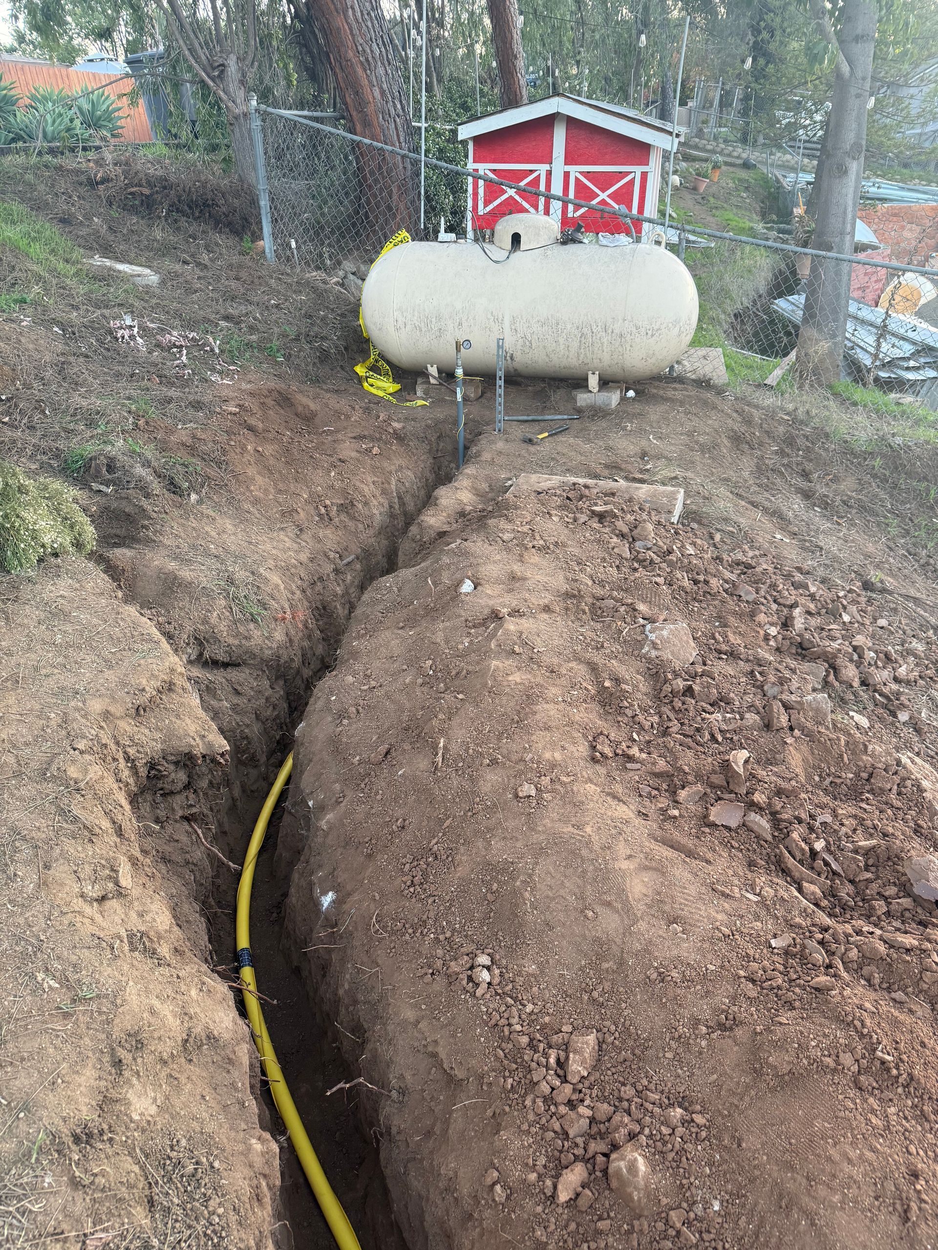 A yellow gas pipe runs through a trench in the ground toward a large, white propane tank in a wooded yard.