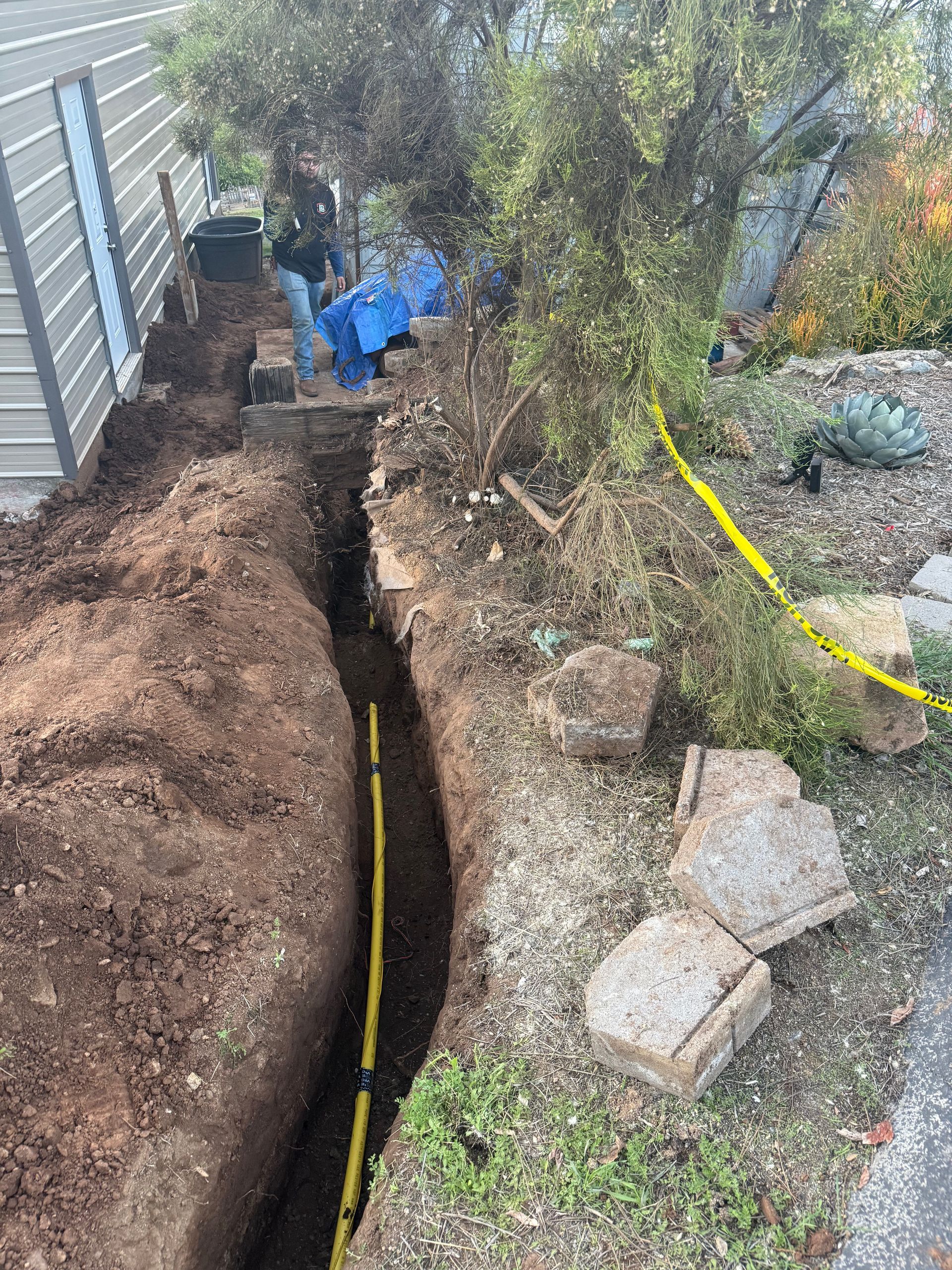 A worker stands in a narrow trench beside a house, where a yellow gas pipe is being installed near a retaining wall.
