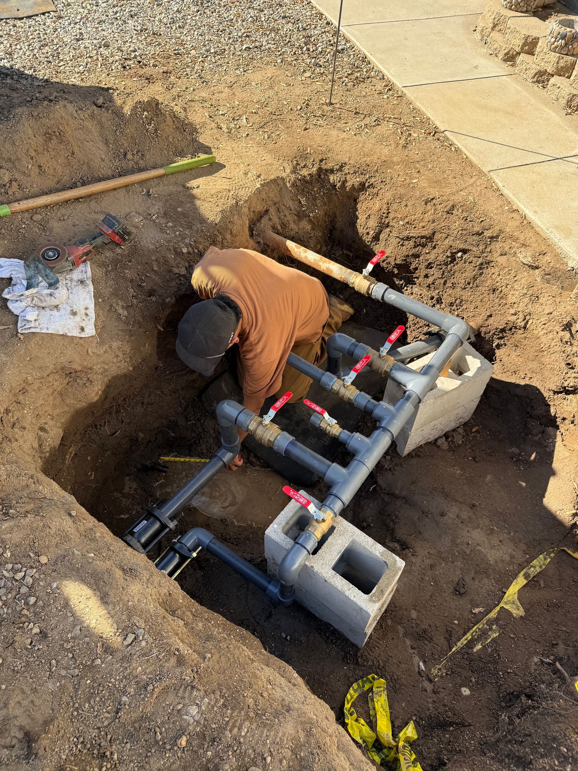 A worker in a brown jacket kneels in a dirt trench, installing a PVC plumbing manifold system supported by cinder blocks.