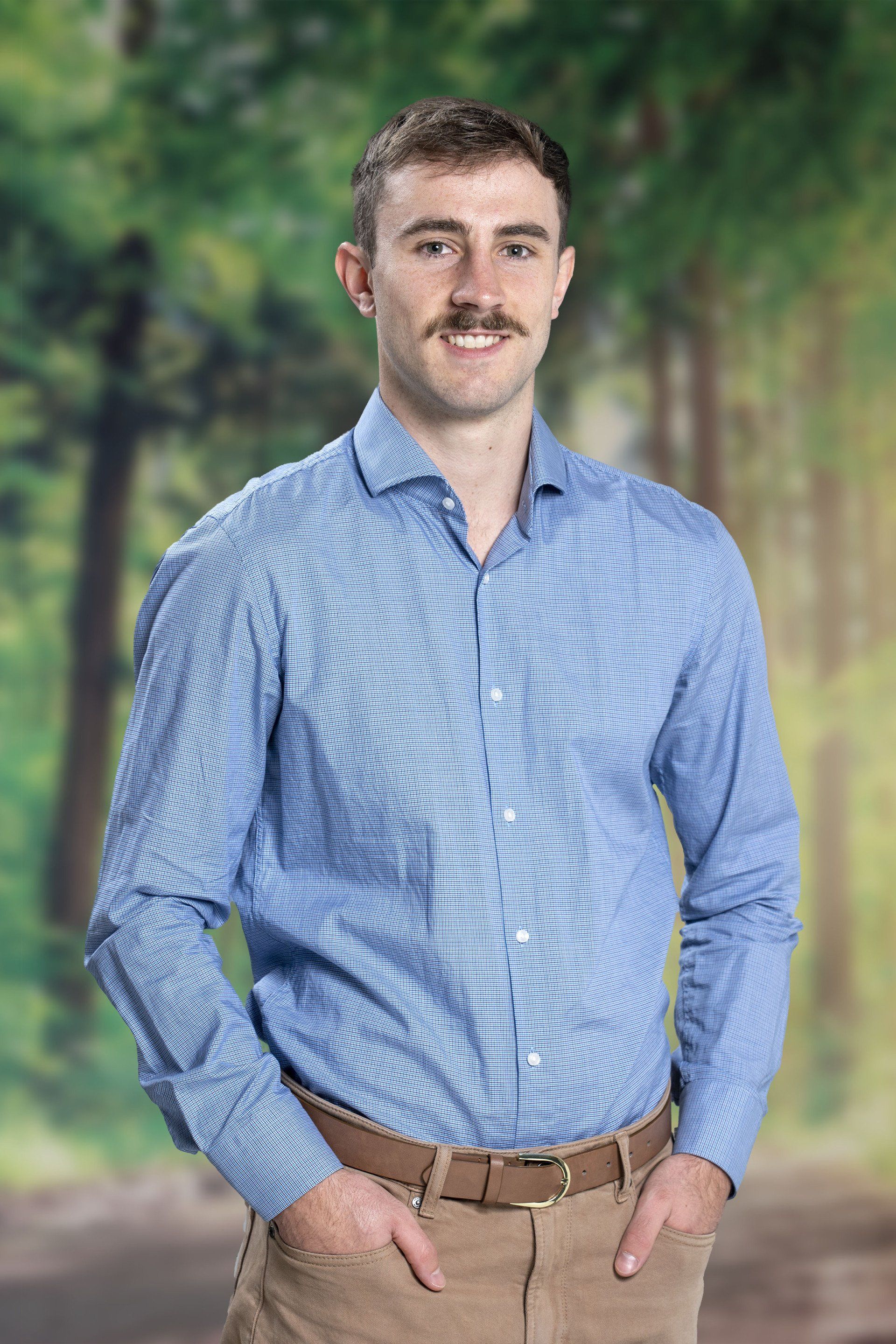 A man in a blue shirt and brown pants is standing with his hands in his pockets.