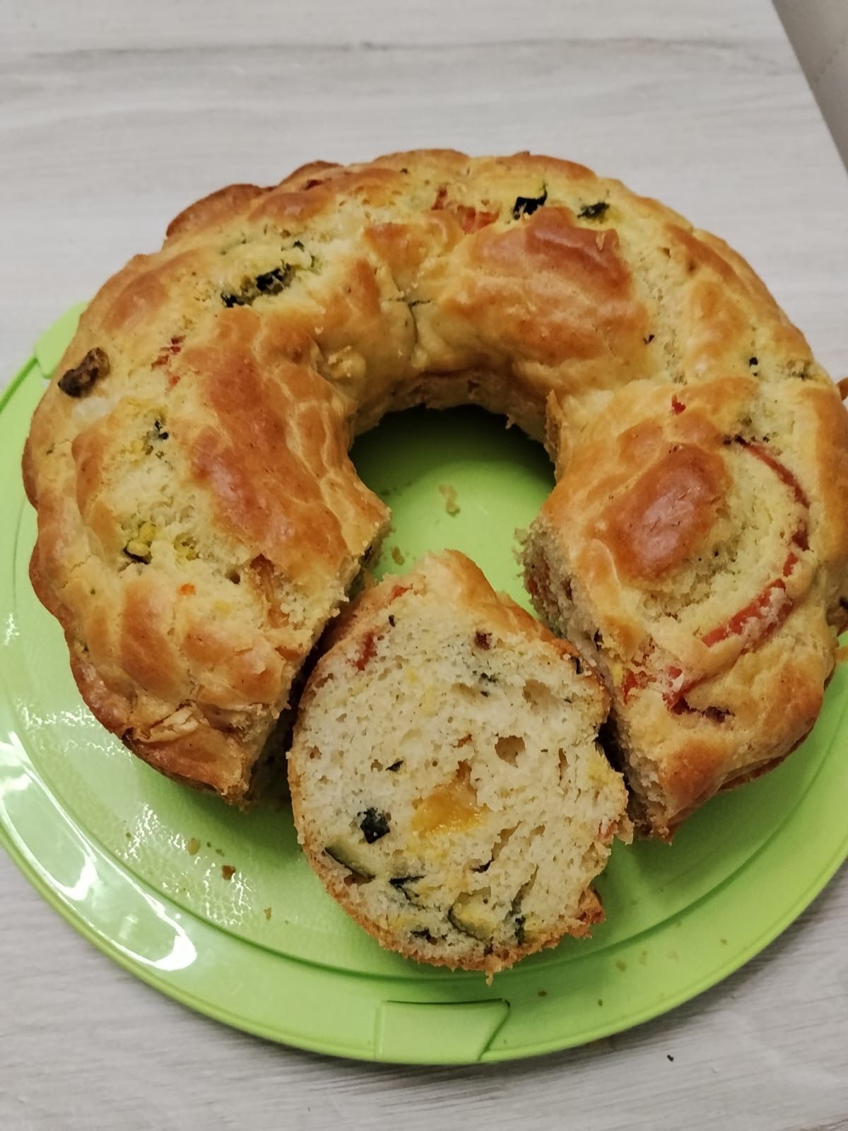 Pagnotta salata a forma di anello con erbe aromatiche su un piatto verde, una fetta tagliata e inclinata in avanti