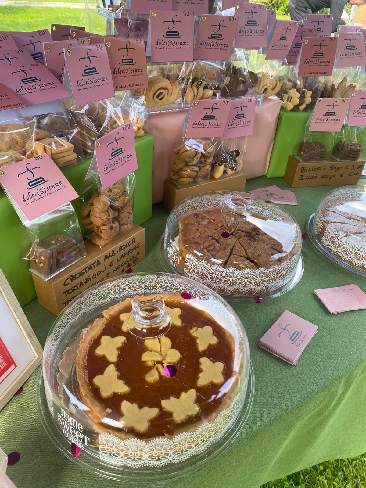 Dolci fatti in casa assortiti su un tavolo verde, con etichette rosa e biscotti, torte e pasticcini confezionati.