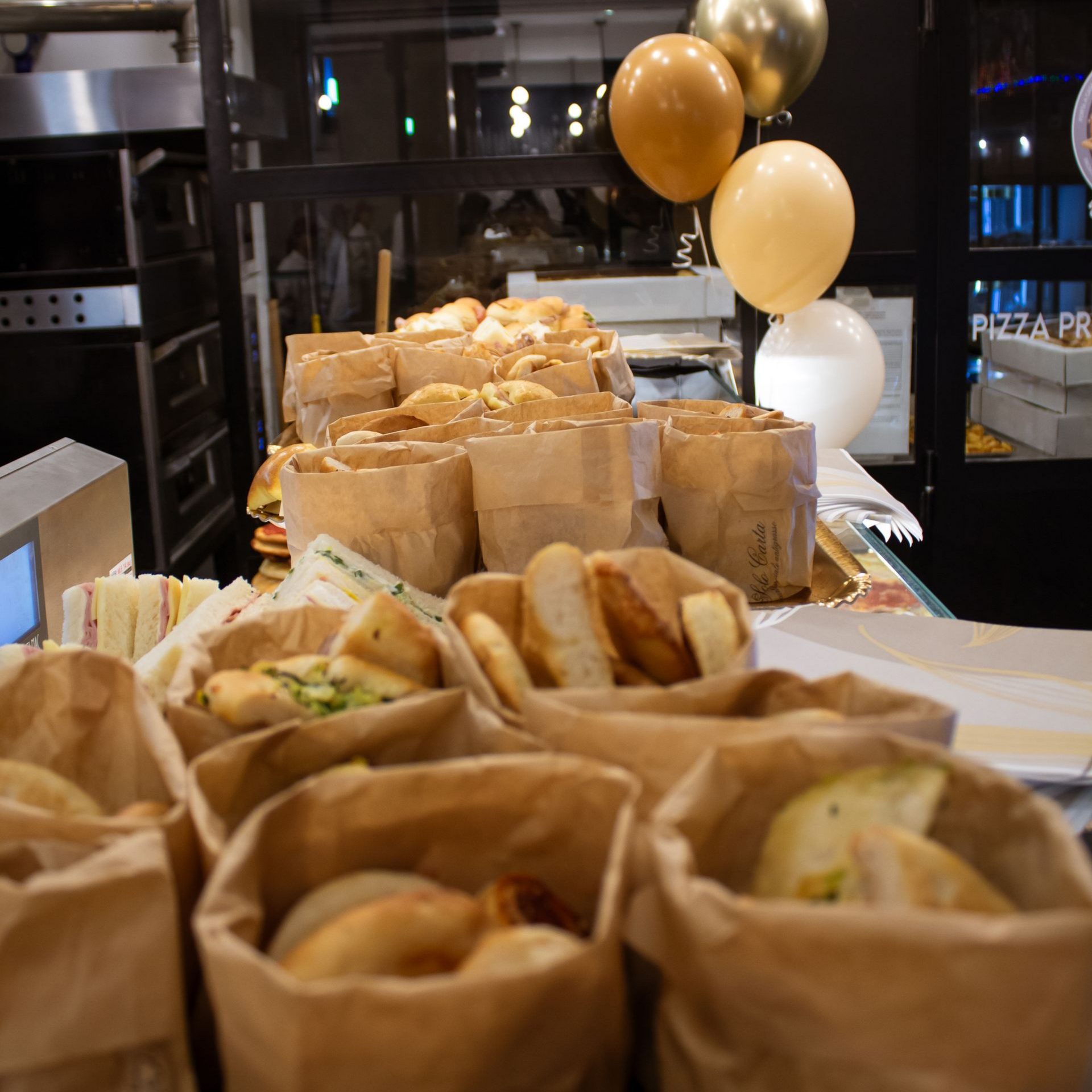 Sacchetti di cibo (panini, pasticcini) su un tavolo, palloncini dorati sullo sfondo.
