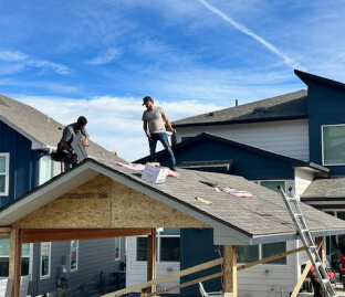 Two workers installing shingles on a house roof beside a ladder and new siding.