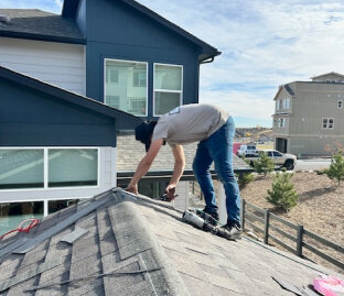 Worker on a roof repairing shingles beside a blue house under a bright sky