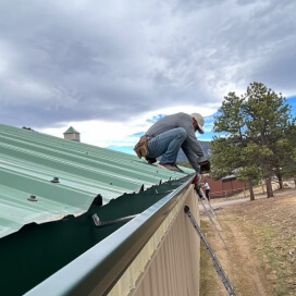 Worker repairing a green metal roof beside a ladder under cloudy skies