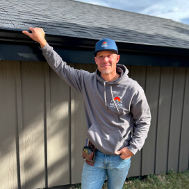 Man in gray hoodie and blue cap leaning against a building roofline, smiling outdoors.