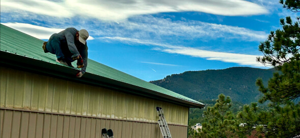 Person crouching on a green roof beside a tan building, with mountains and trees in the background.