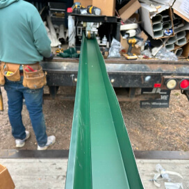Worker guiding a long green metal beam into a truck-mounted machine at a worksite.