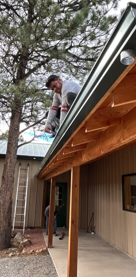 Worker on a roof beside a tan building, inspecting the gutter and soffit near a porch.