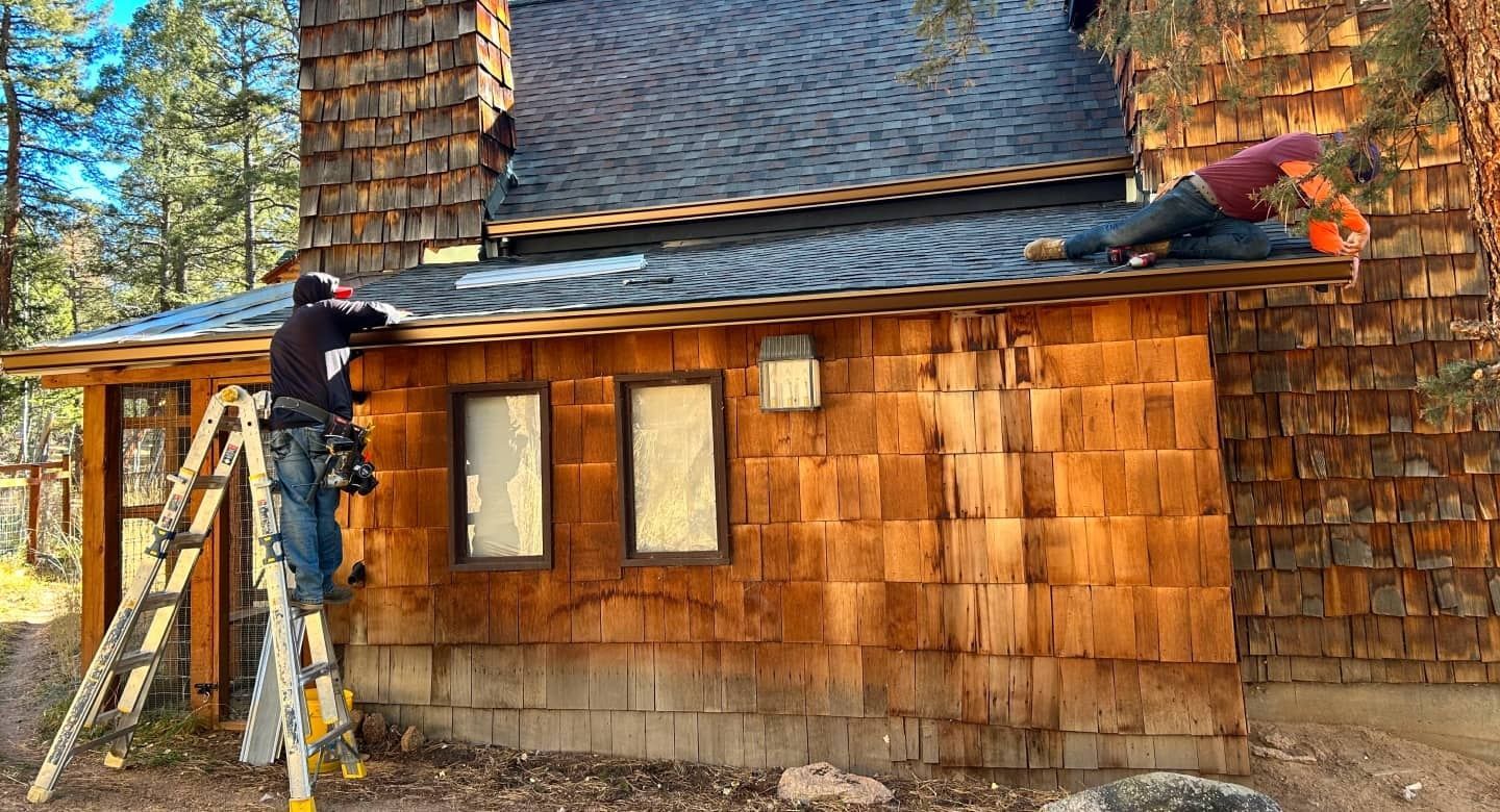 Two workers install wooden shingles on a rustic house exterior, one on a ladder and one on the roof.