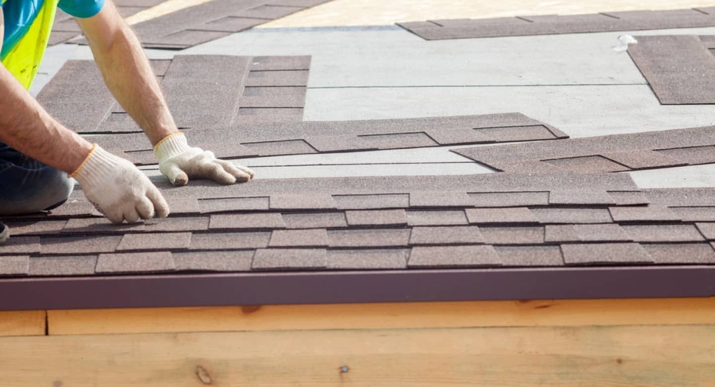 Gloved hands installing brown roof shingles on a house roof