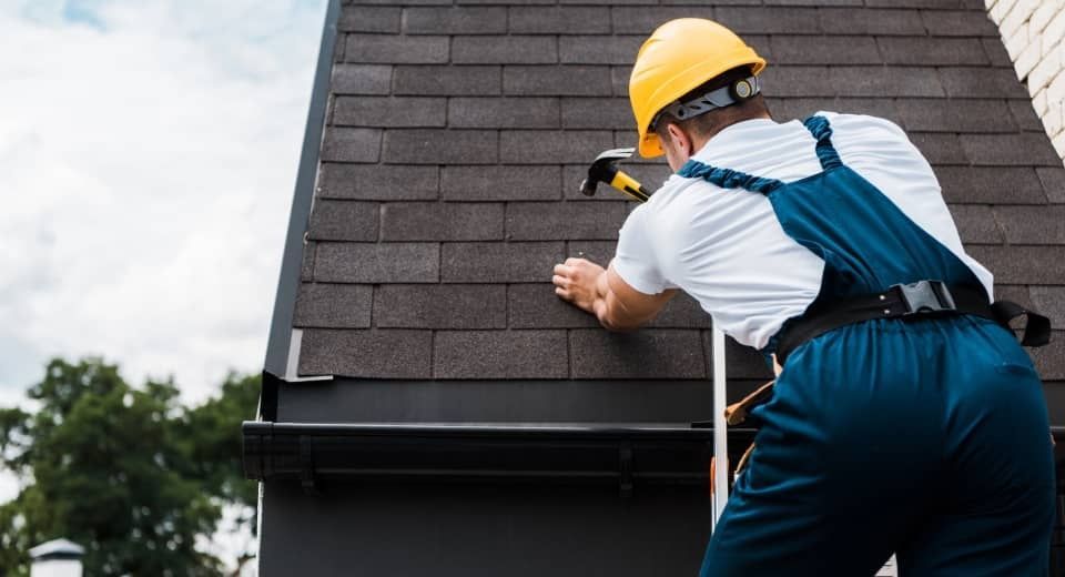 Worker in yellow hard hat inspecting roof shingles on a house with a tape measure.