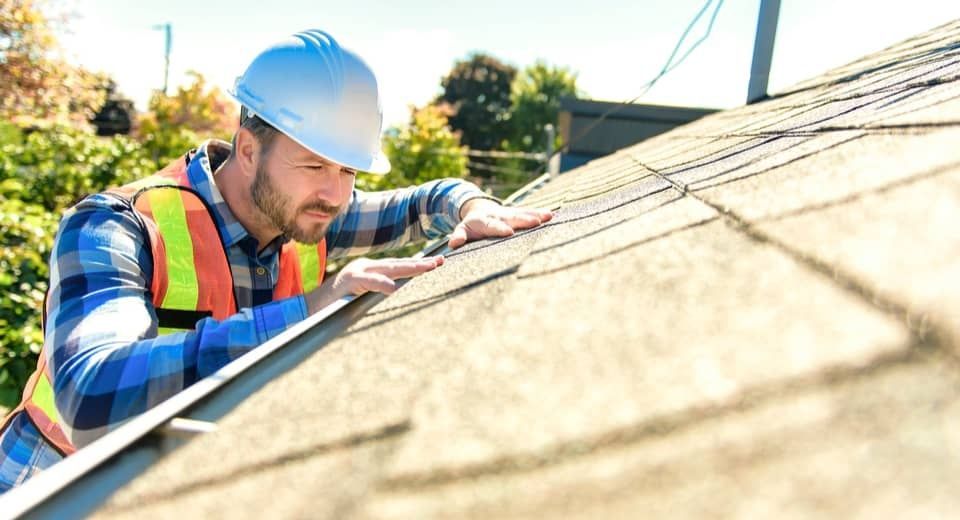 Worker in hard hat and safety vest inspecting a shingled roof with a tool