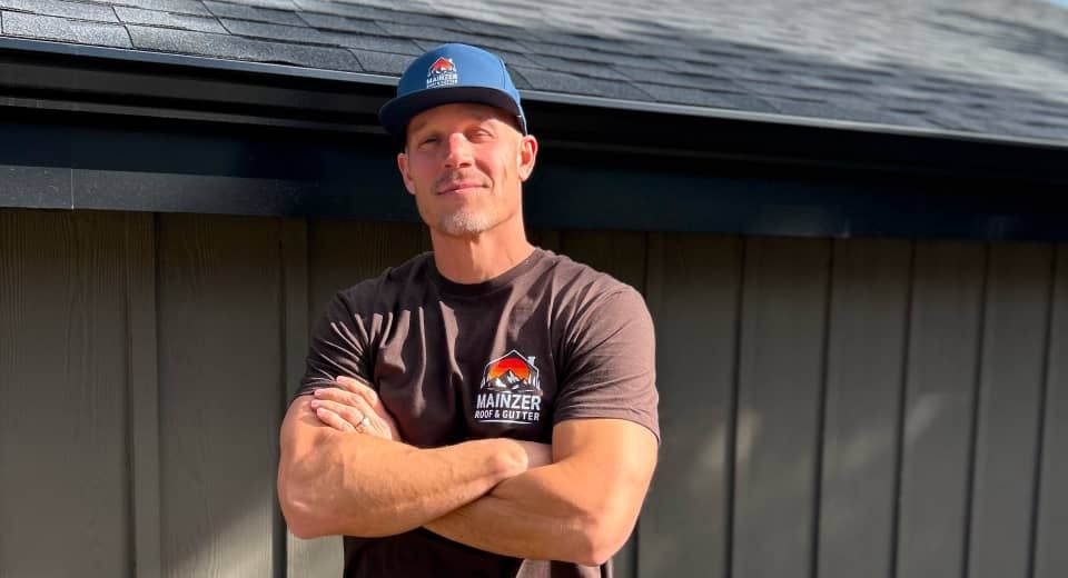 Man standing with arms crossed in a black shirt and blue cap against a building wall