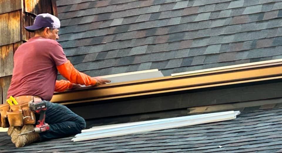Worker installing shingles on a roof, kneeling beside stacked materials and tools