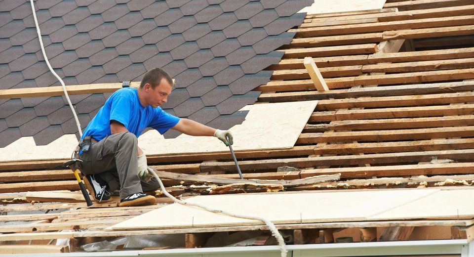 Worker installing roofing on a partially stripped roof, with gray shingles and exposed wooden rafters.