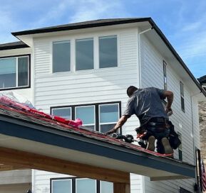 Worker installing roofing on a white house exterior, seen from a low angle
