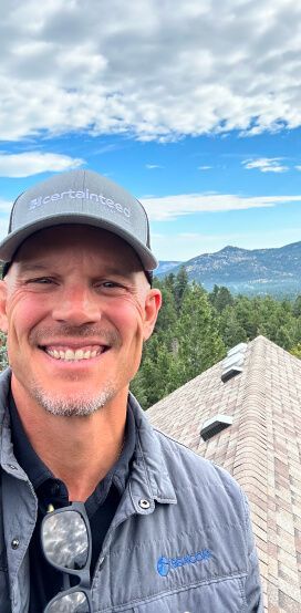 Smiling man in a cap and gray jacket with mountains and blue sky behind him