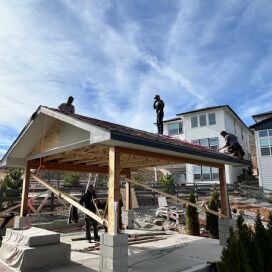 Workers on a rooftop building a pavilion, with lumber, tools, and a blue sky overhead