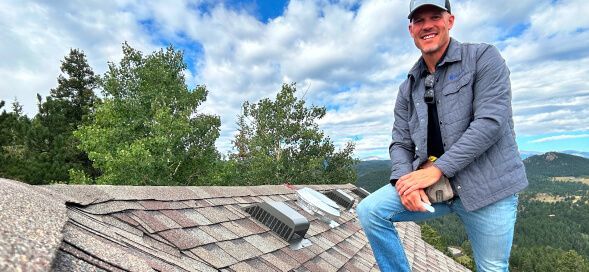 Man sitting on a shingled roof under a cloudy sky, smiling and wearing a cap and blue jacket