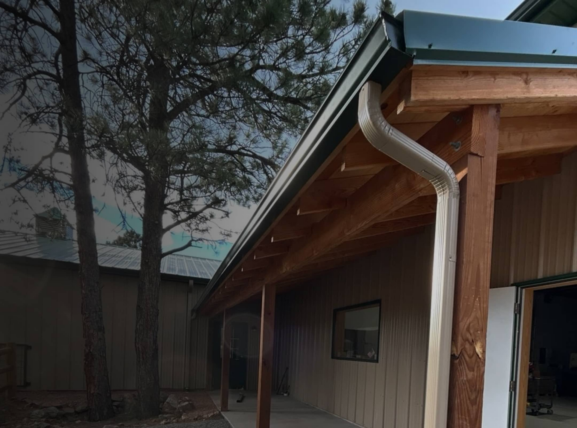 Green rain gutter and light-colored downspout along a timber-framed porch beside pine trees