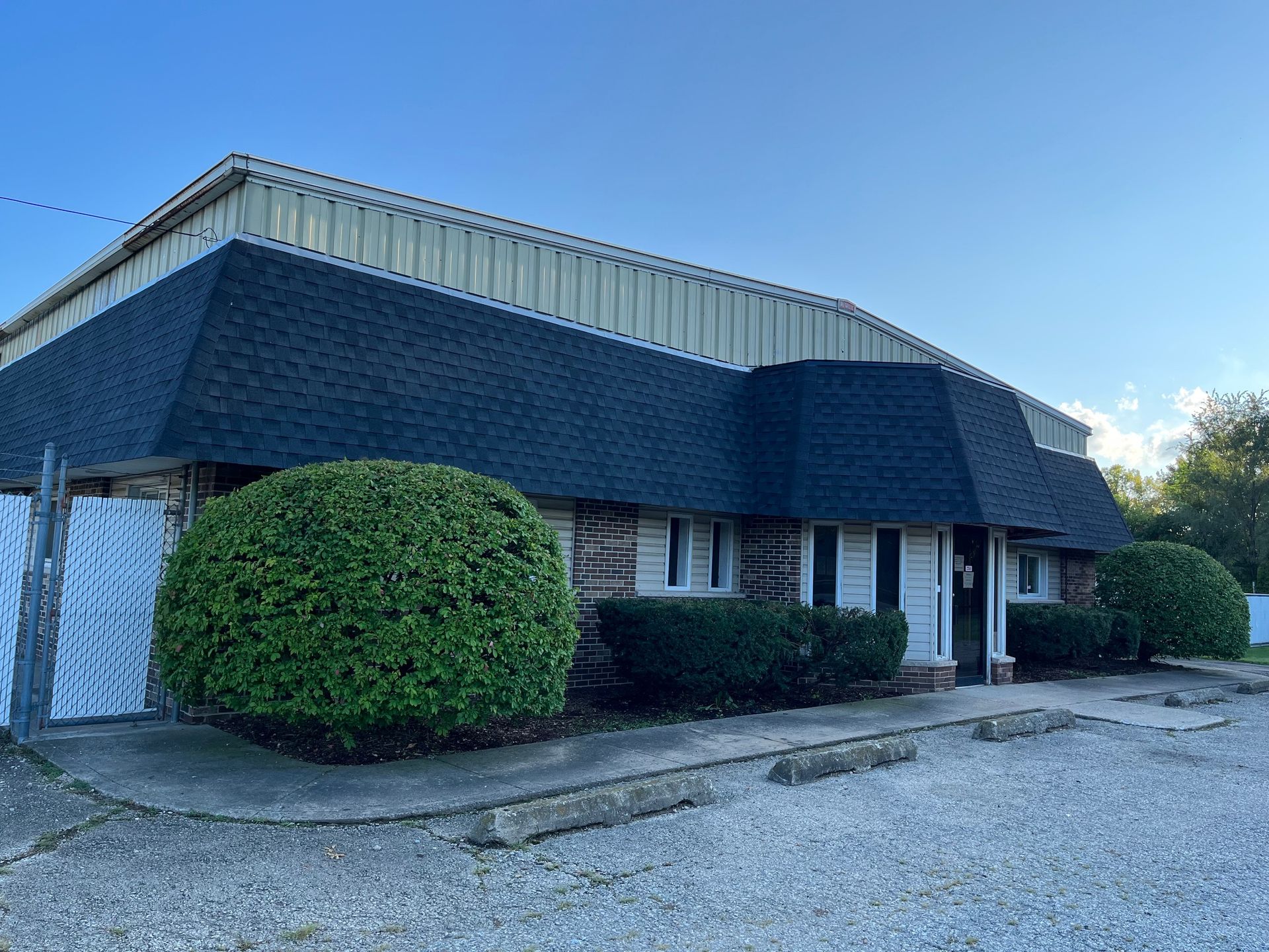 A tan metal-sided industrial building with a brick facade and a gravel parking lot under a clear blue sky.
