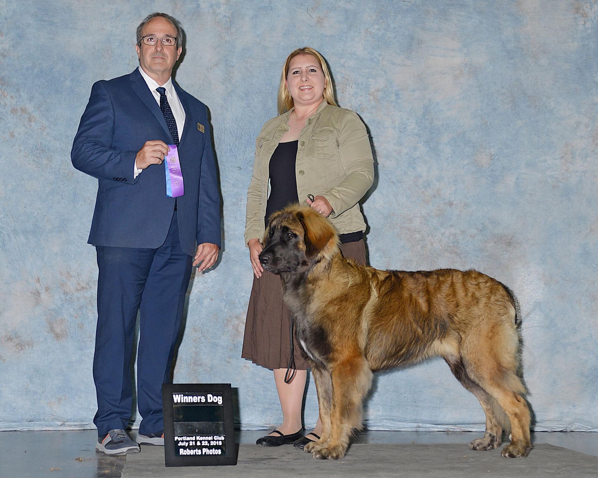 Man in blue suit and woman with large dog posing on a show stage with a trophy plaque