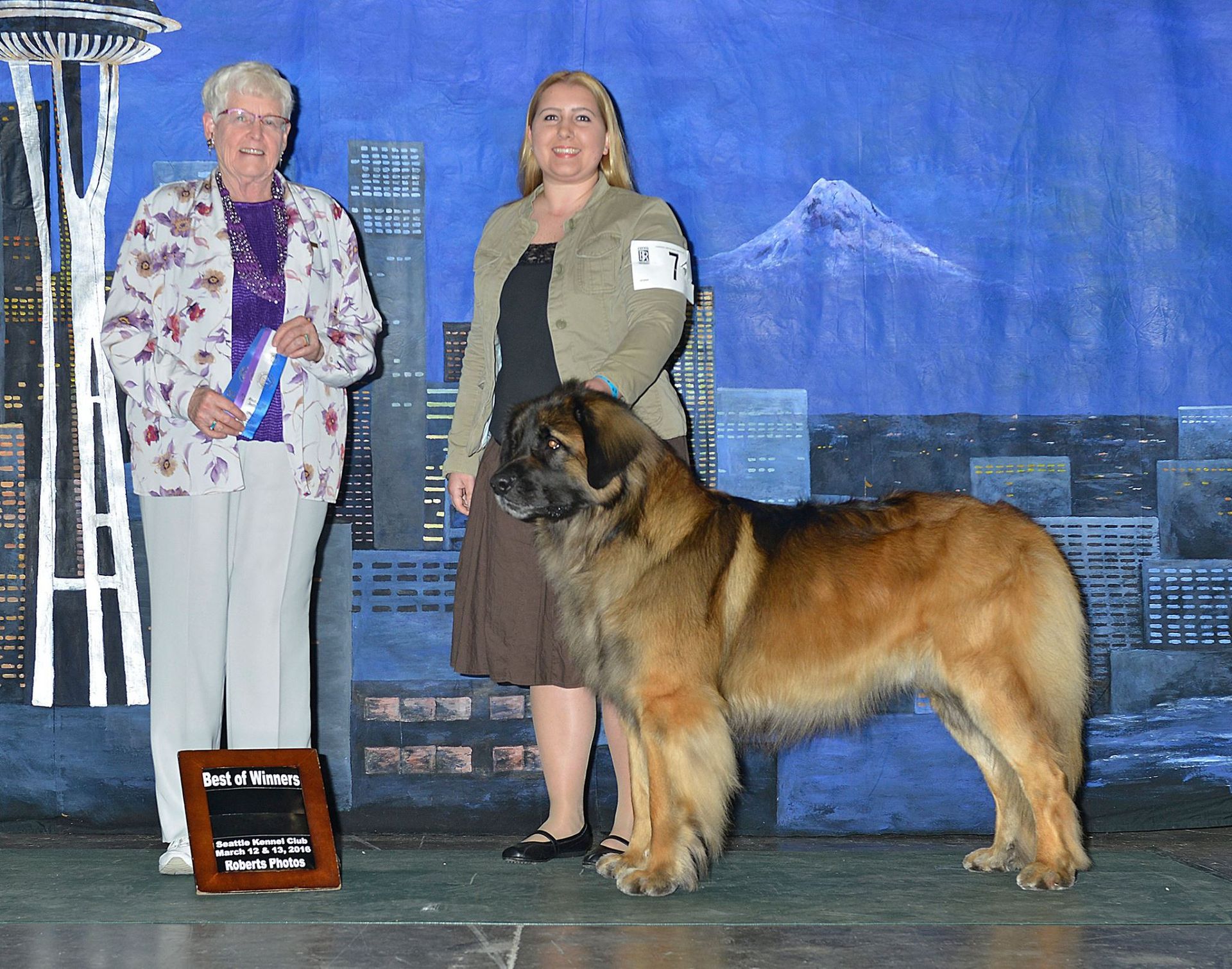 Two women stand with a large tan dog on a stage, with a cityscape backdrop and award sign.