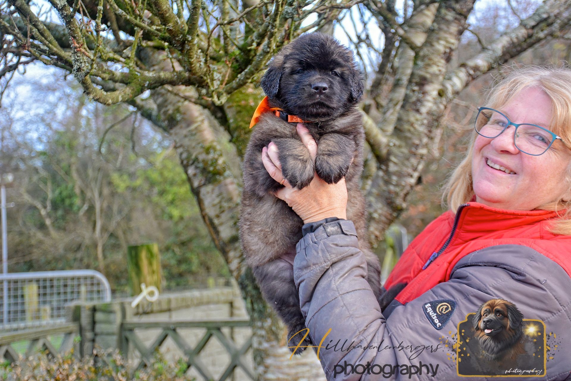 Person holding a small black dog outdoors near a tree and bridge, smiling in a red scarf