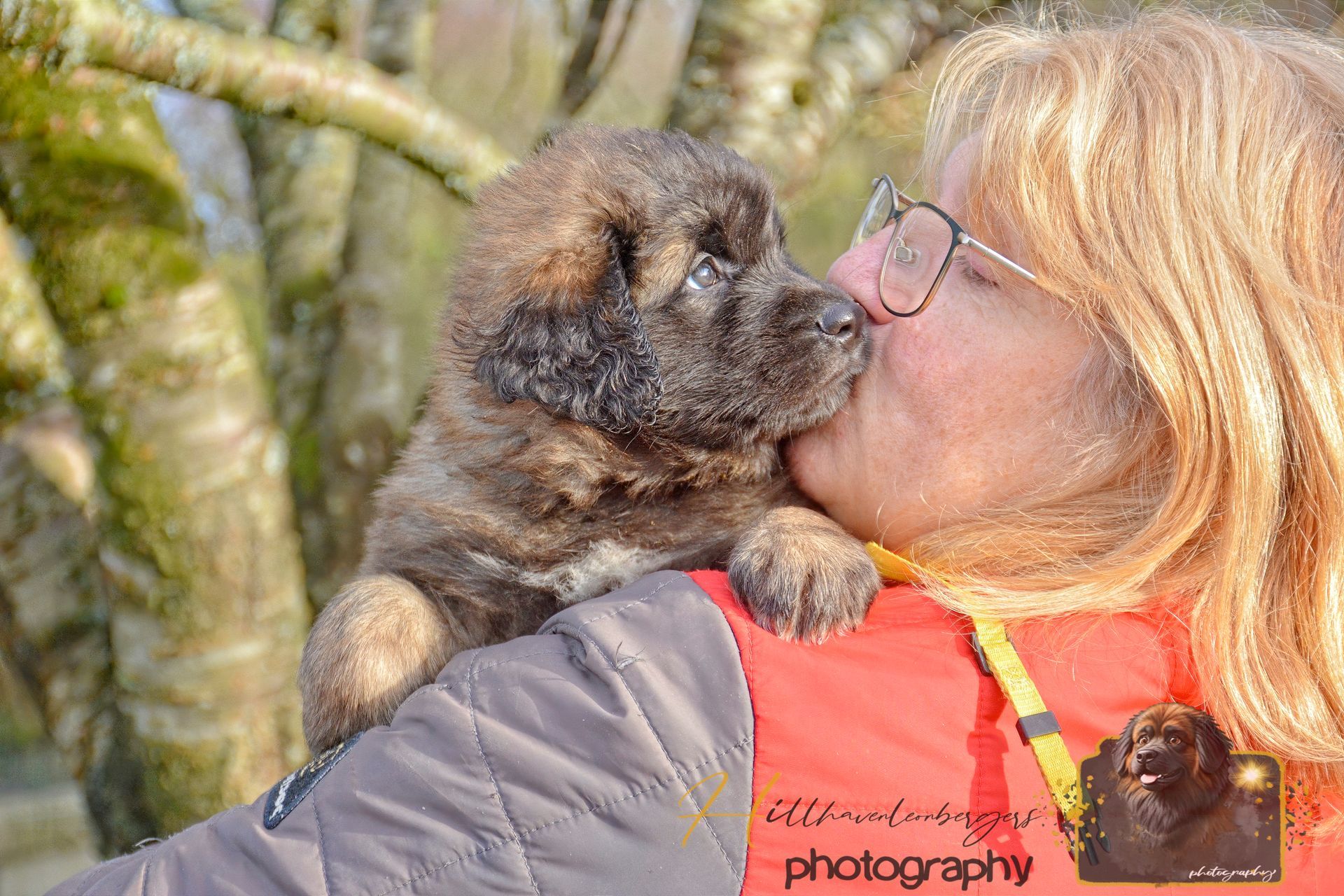Woman in glasses kissing a fluffy puppy outdoors, with trees in the background.