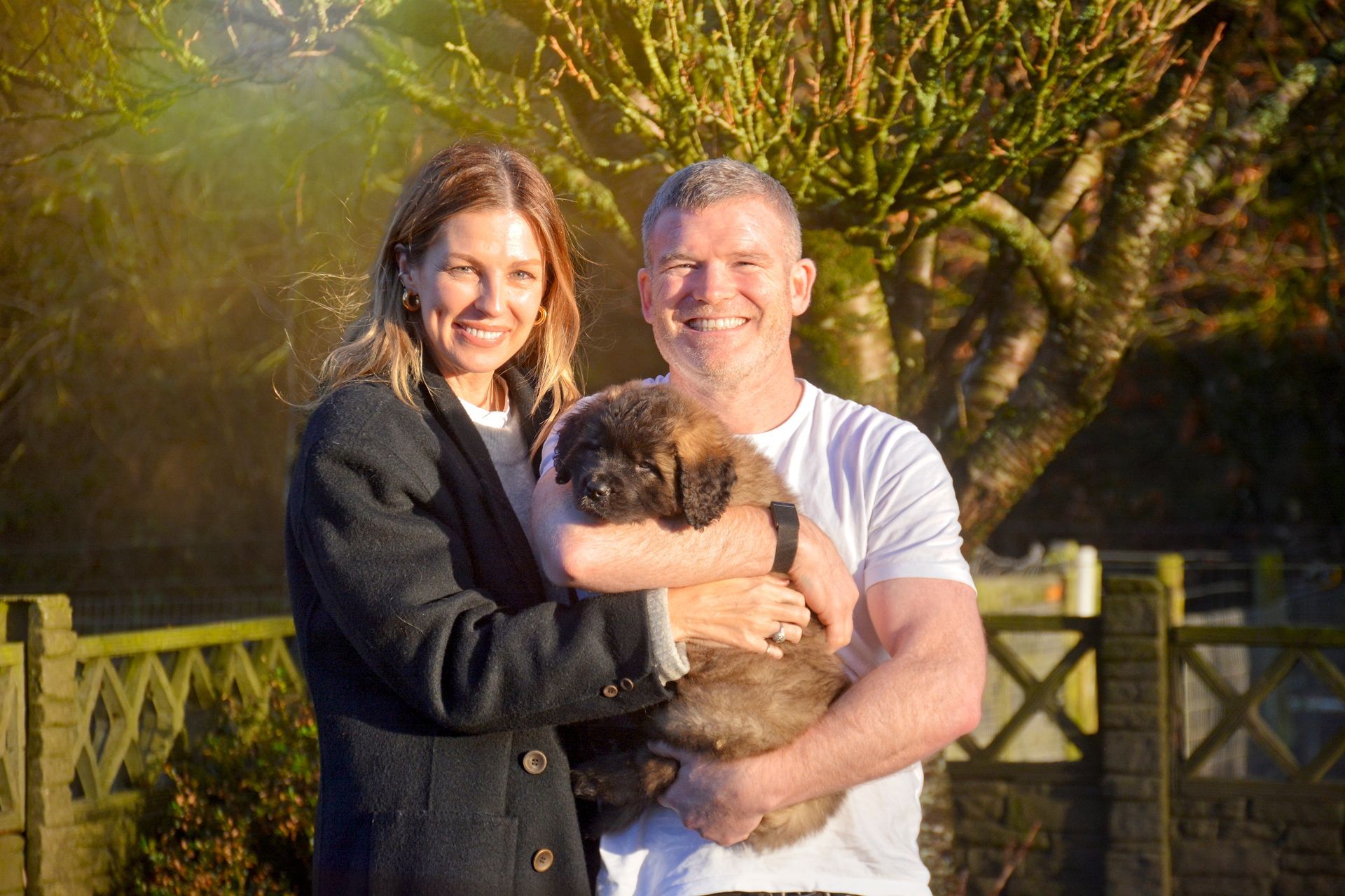 Smiling couple holding a fluffy dog outdoors beside a wooden fence in warm sunlight