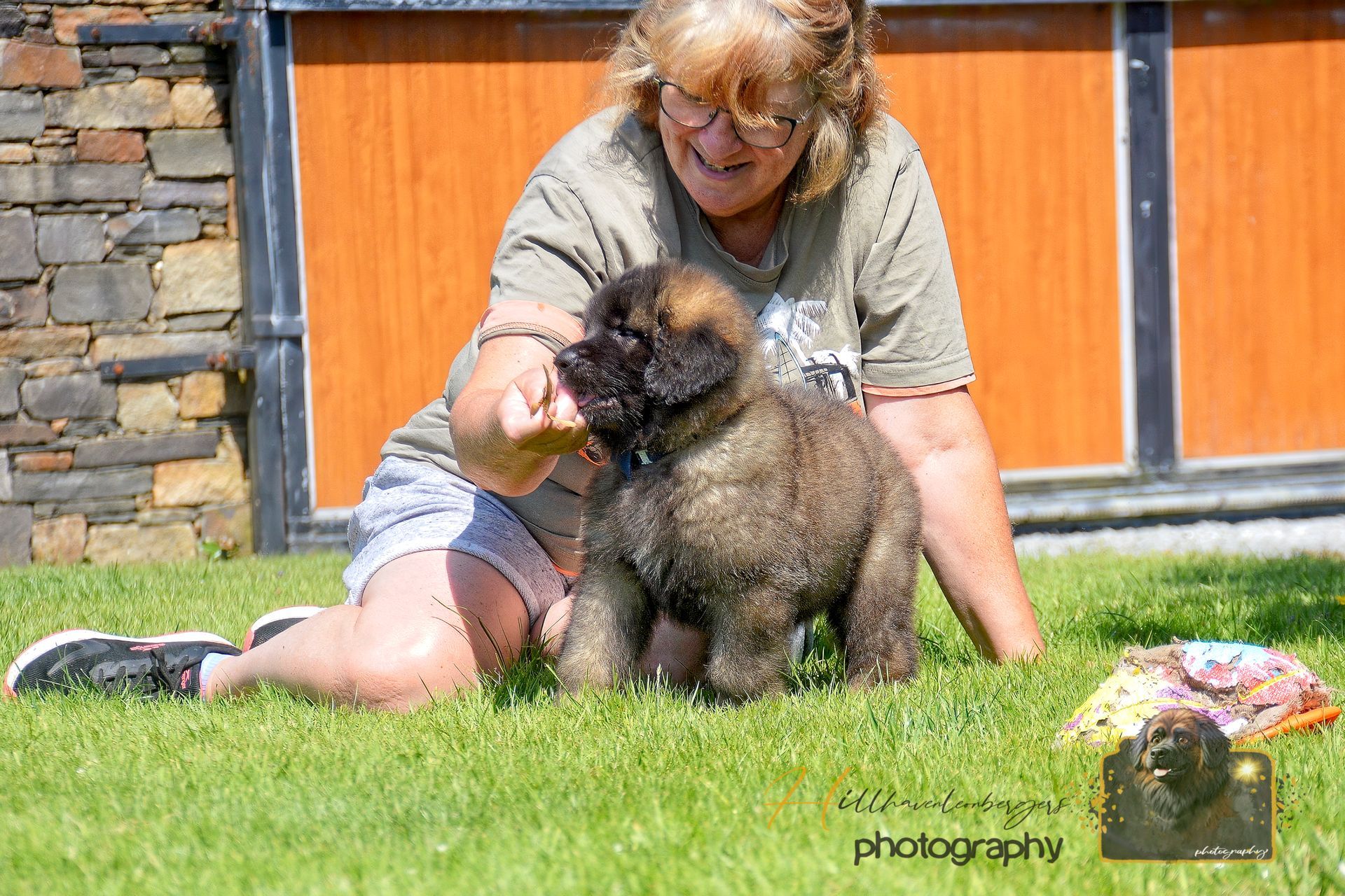 Woman kneeling on grass with a small black dog, petting it outside a stone building.