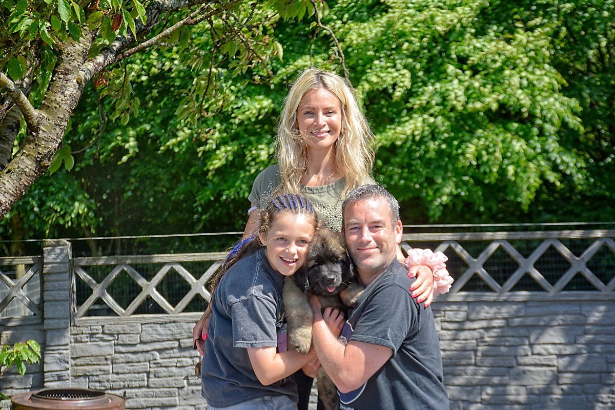 Three people smiling outdoors in front of a stone wall and green trees, two hugging in front of one standing behind