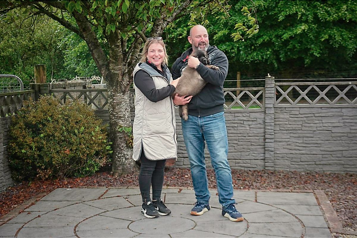 Two people holding a small dog stand on a stone patio by a fence and trees.