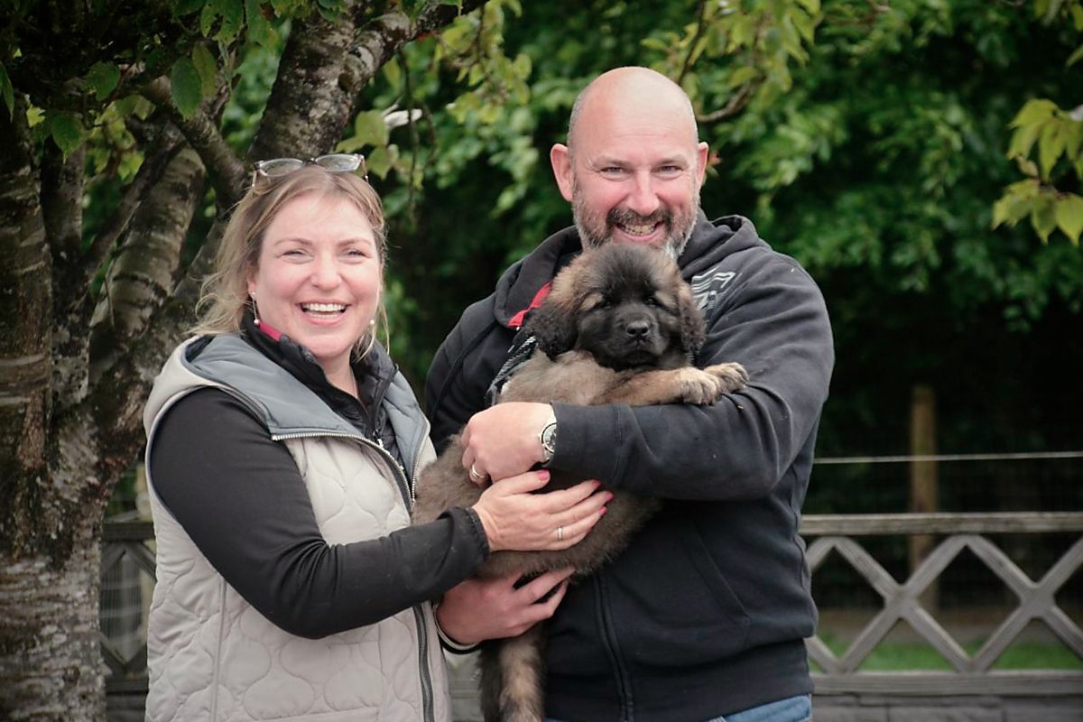 Smiling couple holding a small black dog outdoors in front of green trees and a wooden fence