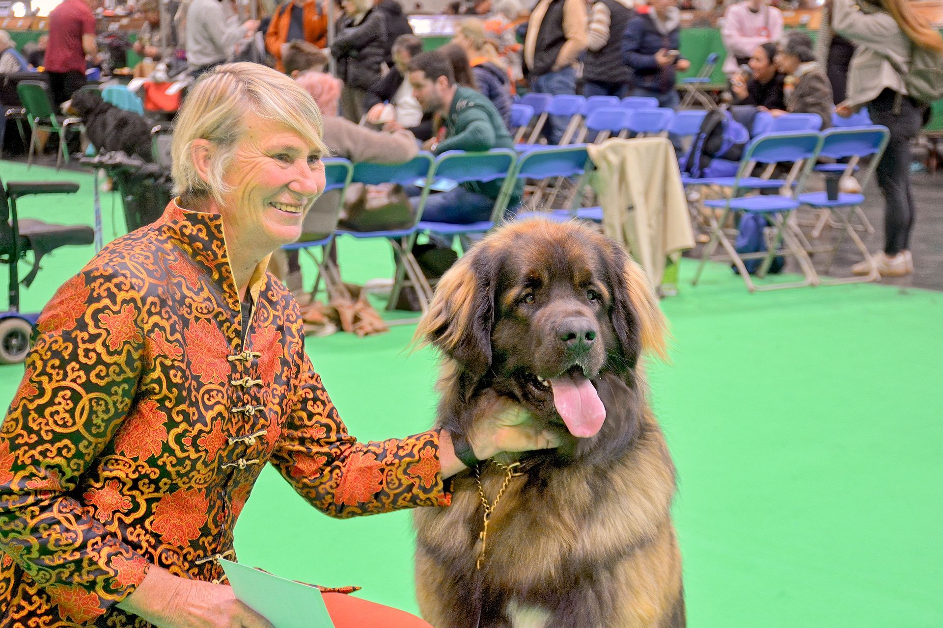 Woman in orange patterned jacket posing with a fluffy dog at an outdoor dog show