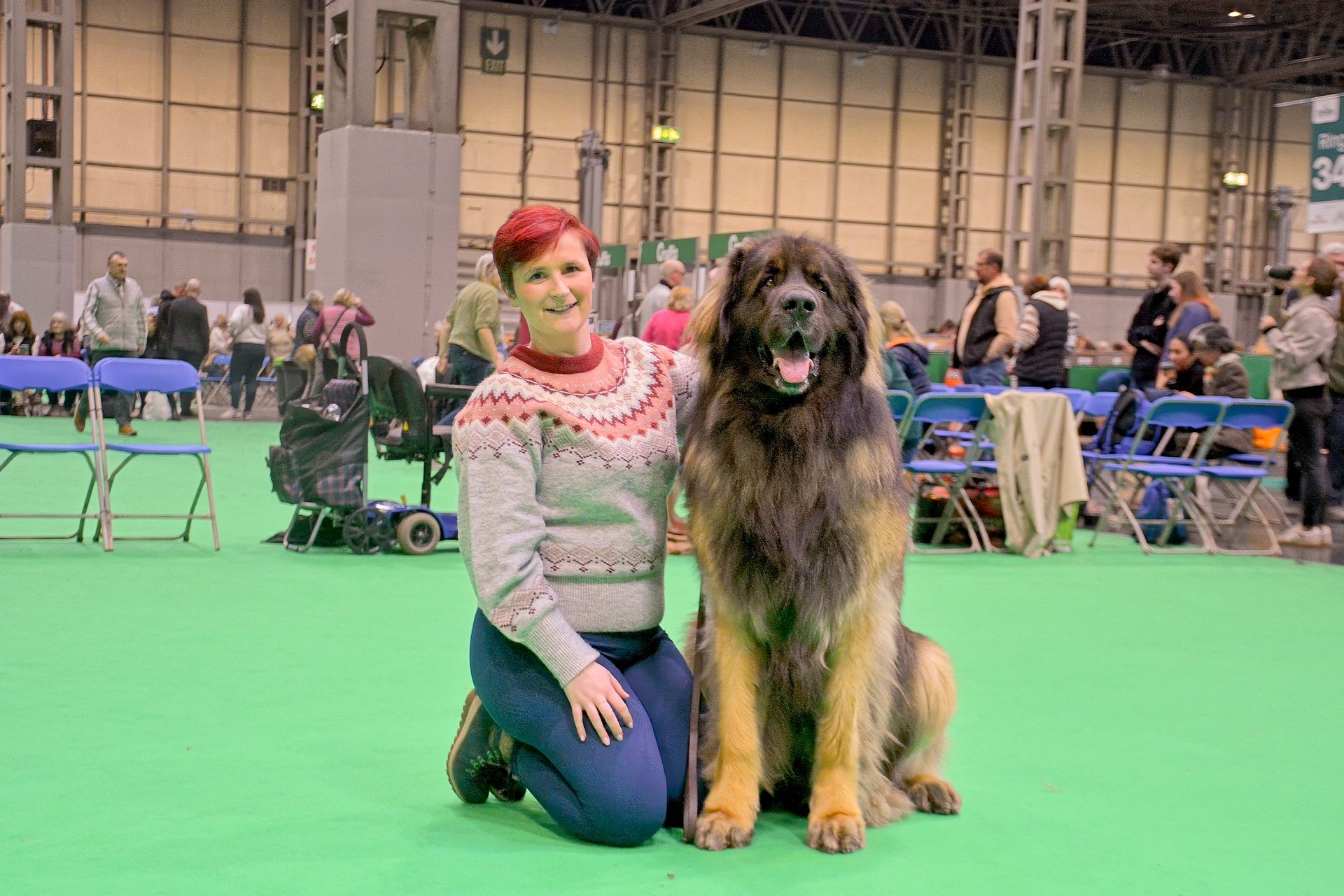 Woman kneeling beside a large fluffy dog at a dog show, with booths and people in the background
